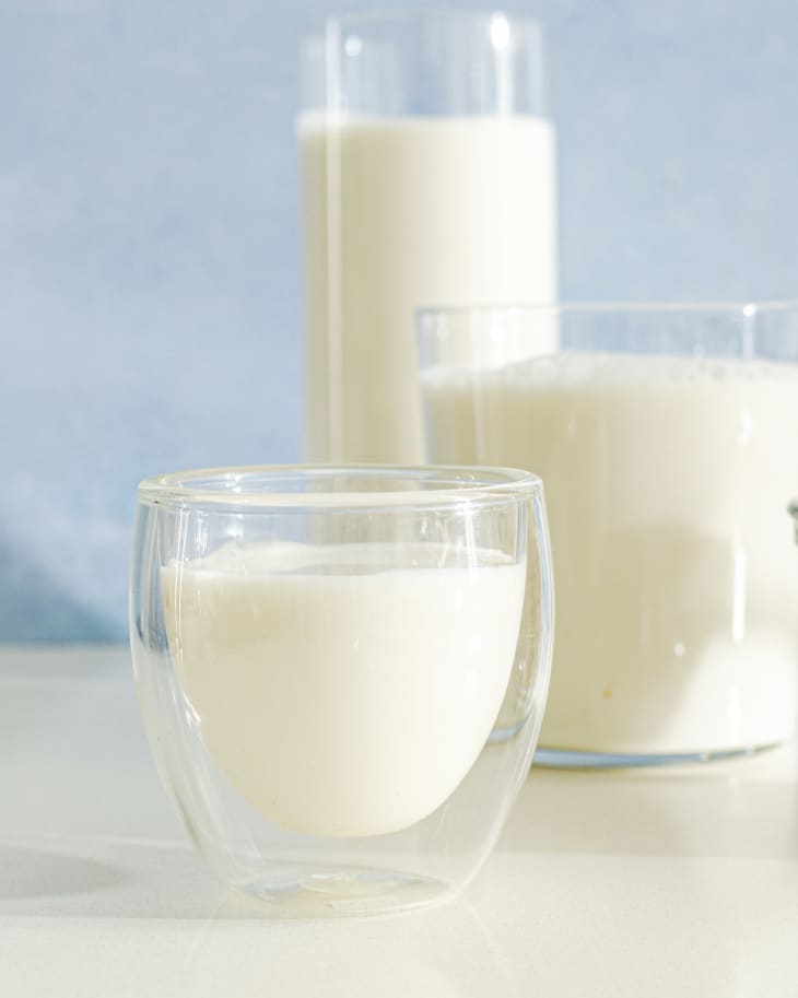 Three glasses of cashew milk (white in color) in glassware of staggered sizes, against a blue background