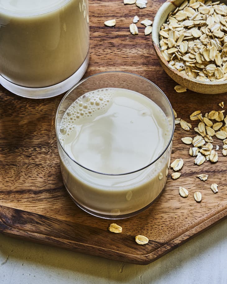 A glass of oat milk on a wooden tray with a bowl of oats in the background