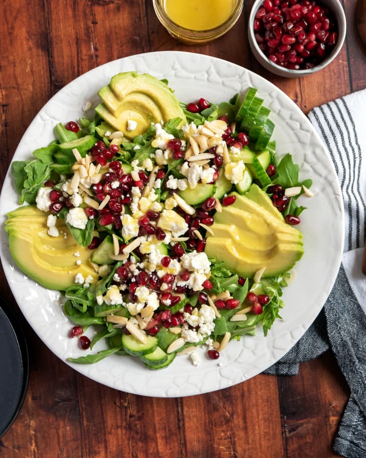 Salad with pomegranate, cheese, nuts and avocado on top, with salad dressing and pomegranate seeds in small bowls nearby