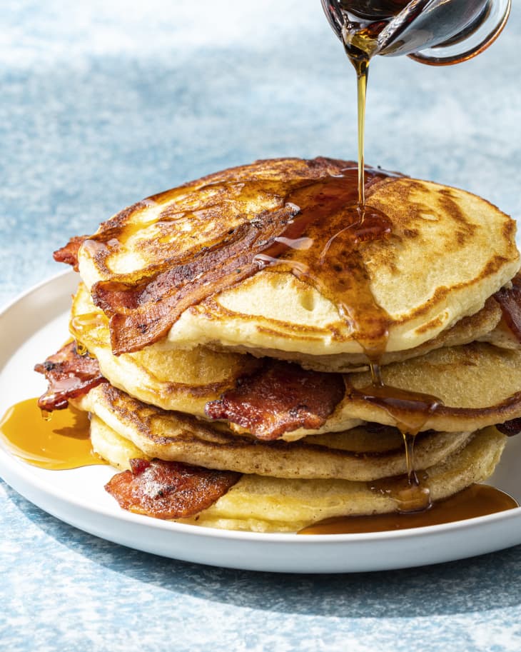pancakes with a slice of bacon cooked into the center, on a round white plate, with syrup being poured over the top.