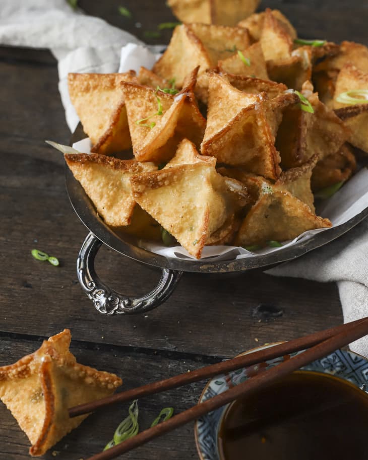 cream cheese wontons that are fried and crispy, in a serving platter with a napkin below, with a bowl of dipping sauce on the side and a pair of chopsticks resting on top.