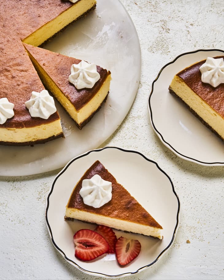 New York style cheesecake  with small dollops of whipped cream on a round serving platter, with two pieces cut out each on their own small dessert plate.