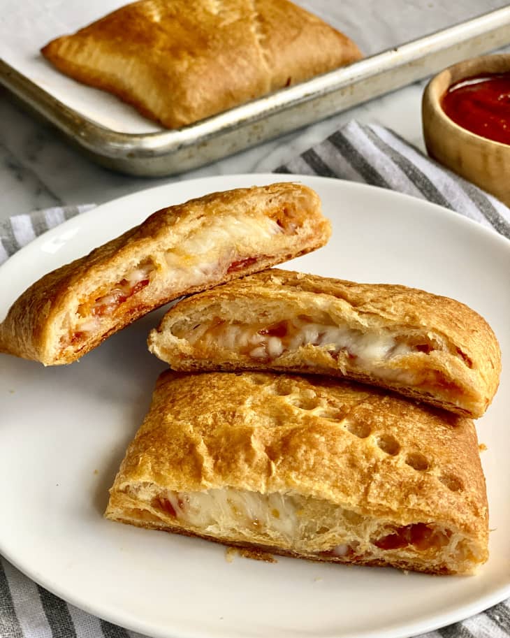 three pizza pockets (dough pockets with pizza fillings inside) on a round, white plate with sauce in a small wooden ramekin next to the plate and another pizza pocket on a square silver baking dish in the background.