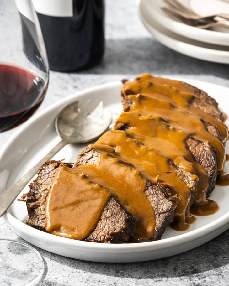 Sauerbraten (a traditional German roast of heavily marinated meat. It is regarded as a national dish of Germany, and is frequently served in German-style restaurants internationally. It can be prepared from a variety of meats, most often from beef) on a white plate with a a spoon resting on it, and glass of red wine and a bottle in the background