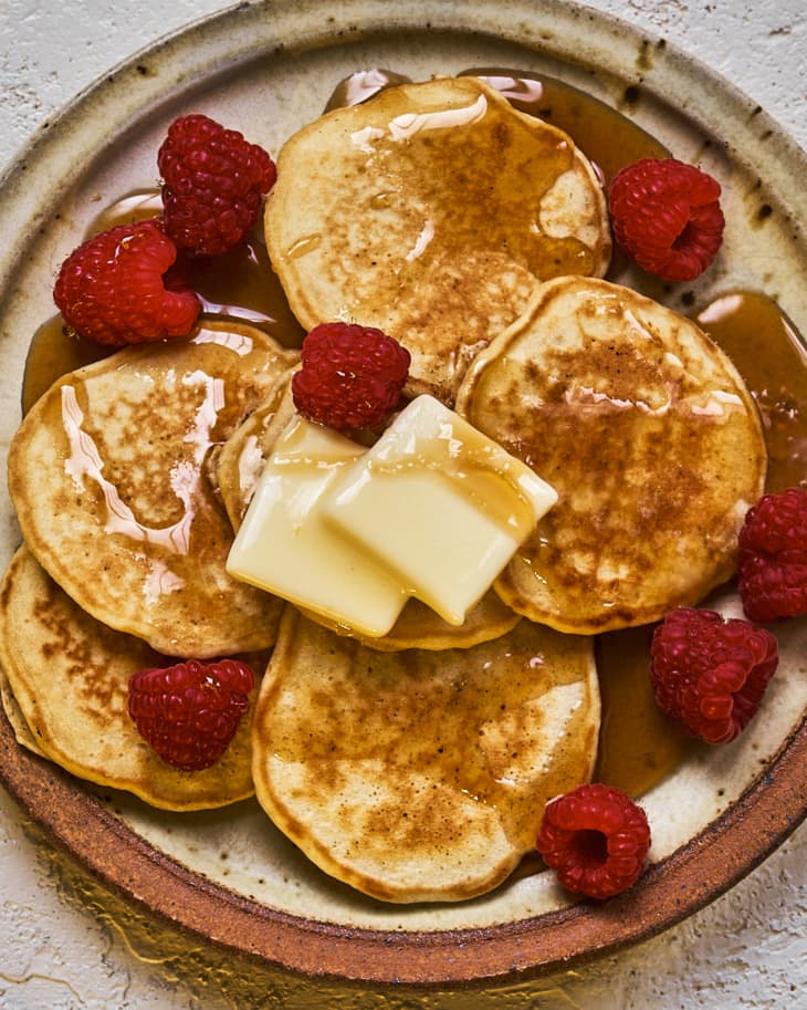Silver dollar pancakes ona. plate with two pats of butter, raspberries and maple syrup drizzled on top.