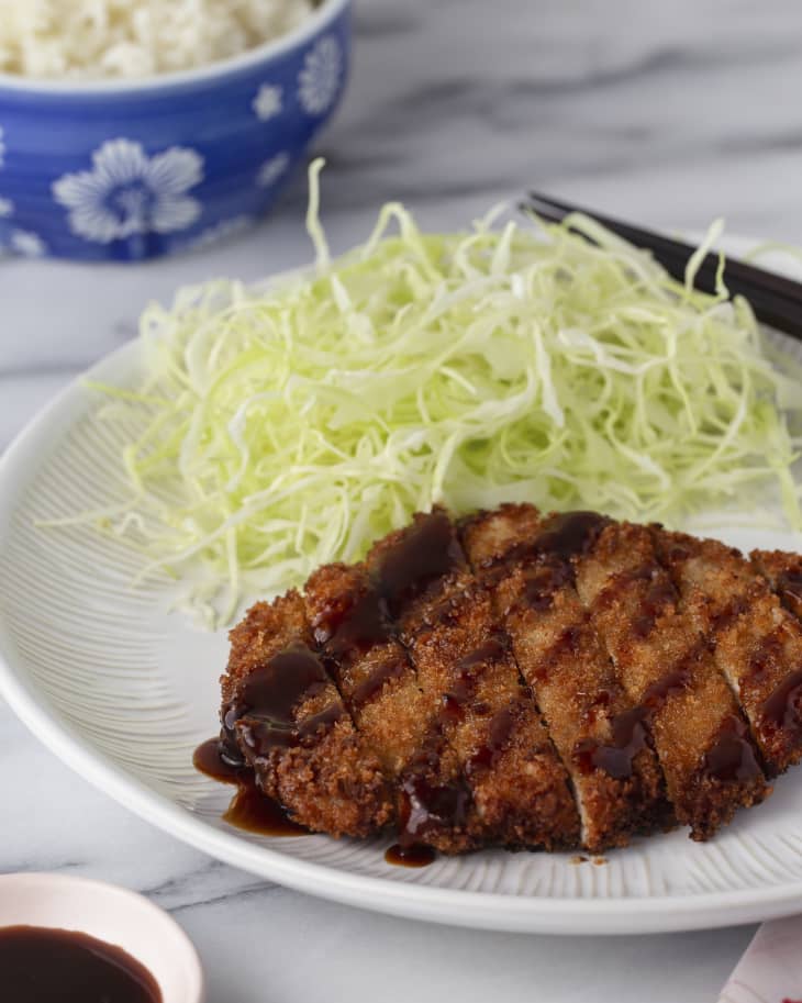 A plate of Tonkatsu (a Japanese dish that consists of a breaded, deep-fried pork cutlet.) It is on a white plate with a side of shredded cabbage on the plate and a bowl of white rice on the side.