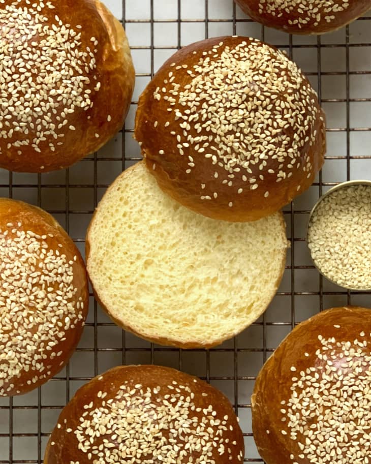 a round sesame seed bun cut in half with other buns around it on a cooling rack.