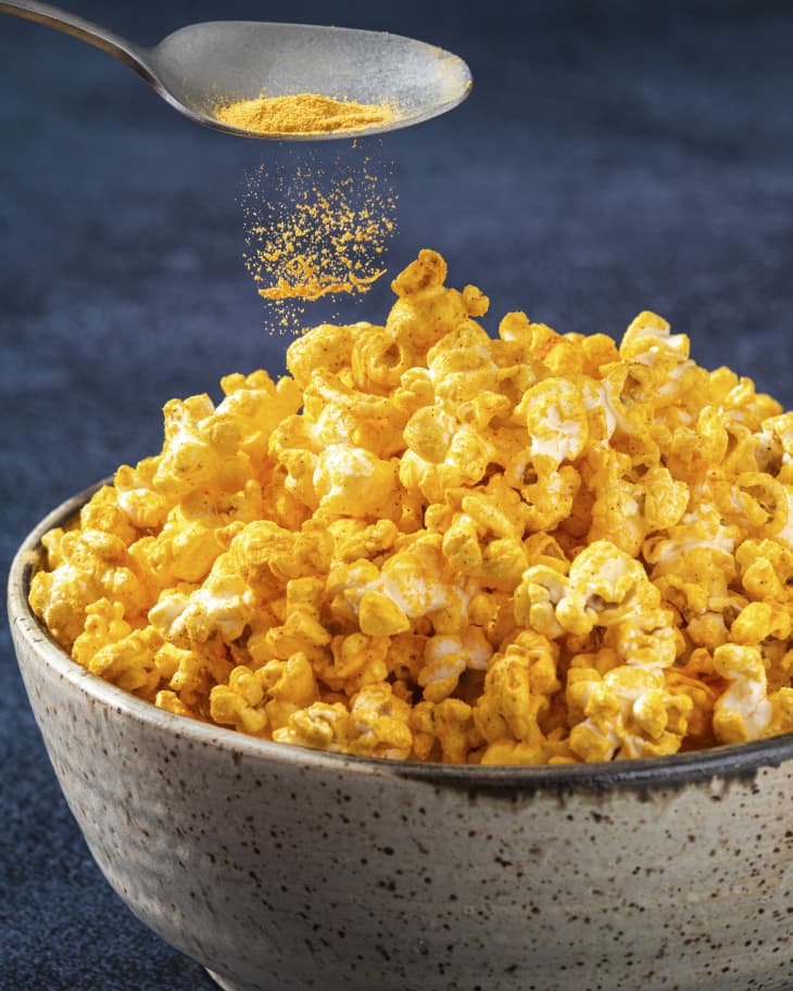 cheesy popcorn in a bowl with a spoon sprinkling more cheese powder over the top.