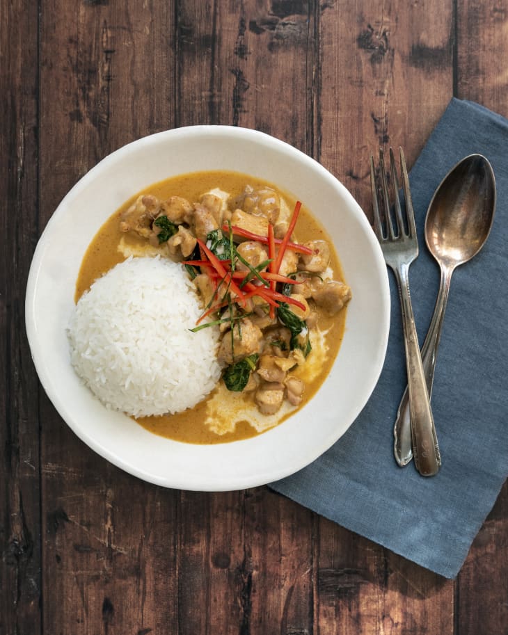 Panang curry (Panang curry (a type of red Thai curry that is thick, salty and sweet) in a bowl with a side of white rice and a fork and spoon resting on a blue napkin on the side.