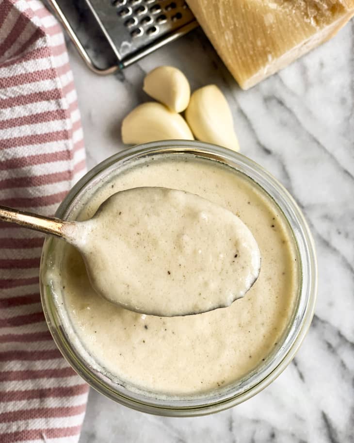 a creamy, white, garlic parmesan sauce in a small glass jar as seen from the top, with pepper mixed in, and a metal spoon lifting some sauce up. There are some garlic cloves next to the jar.