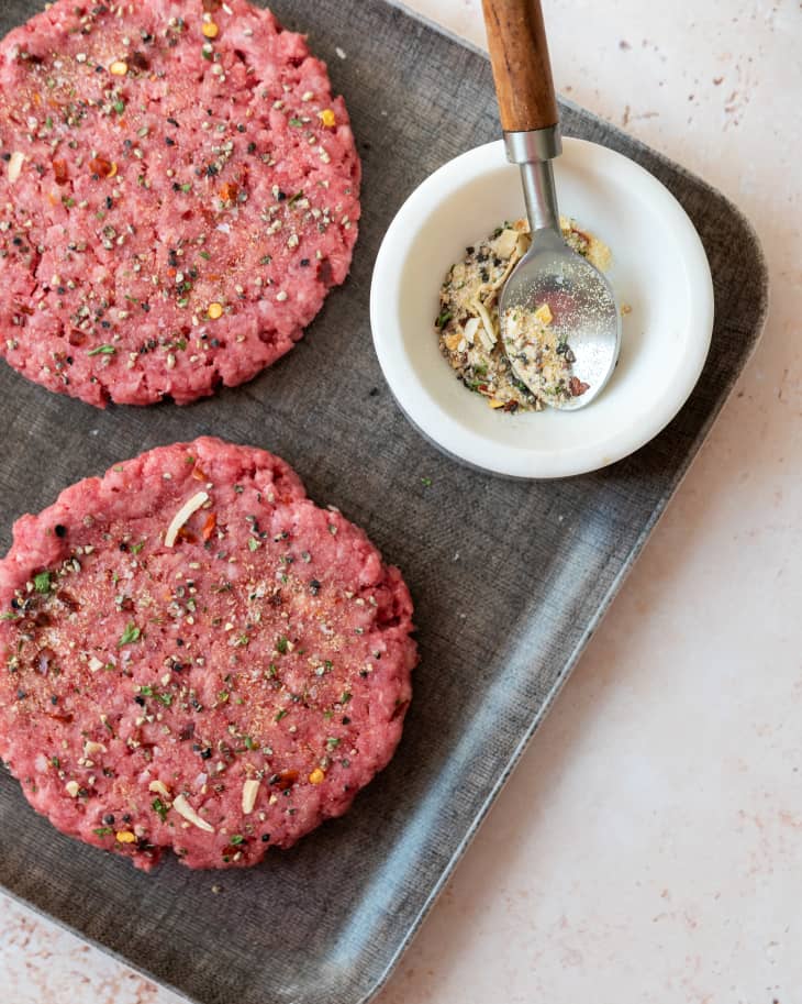 two raw burger patties with seasoning sprinkled on top, and a small bowl with burger seasoning on the side with a silver spoon with a wooden handle resting inside the bowl.
