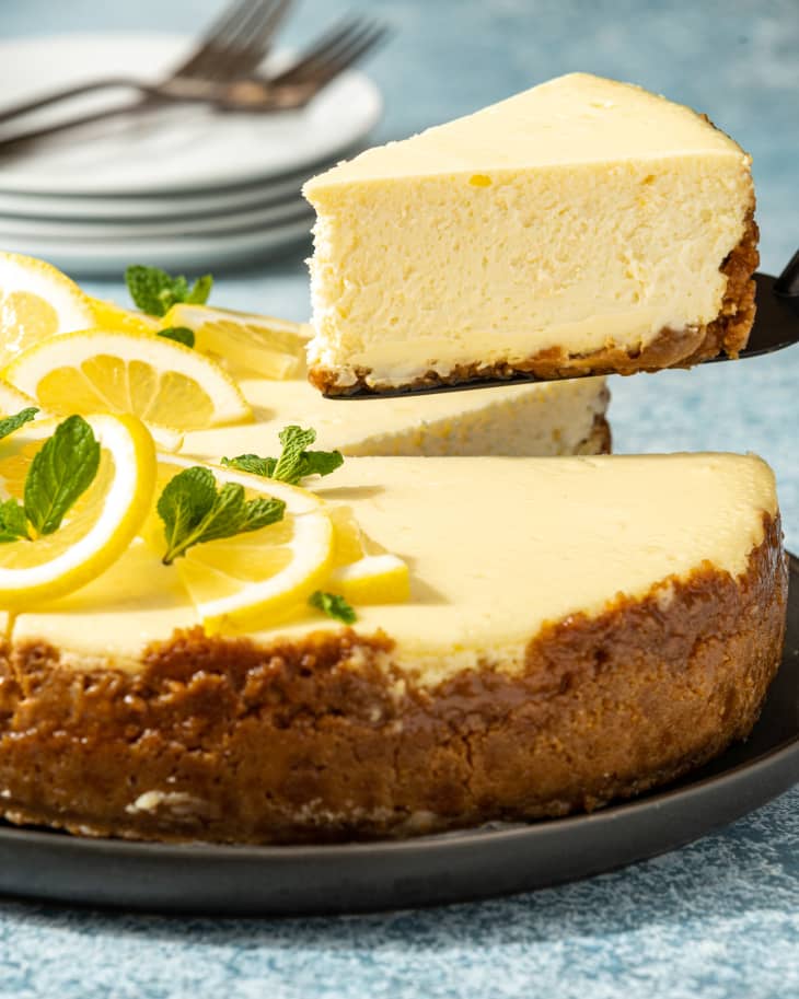 A whole lemon cheesecake with ribbon cuttings of fresh lemon as a garnish on top, with a slice cut out, with the slice being lifted up