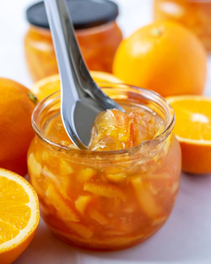 orange marmalade in a small rounded glass jar with a silver scooping spoon starting to spoon a small amount out, with oranges surrounding it