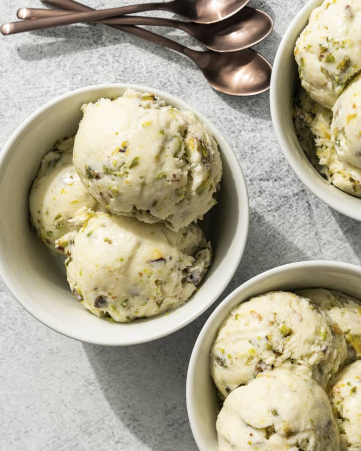 scoops of pistachio ice cream in a white bowl, with two other bowls with the same kind of ice cream next to it, and three silver spoons lying beside the bowls.