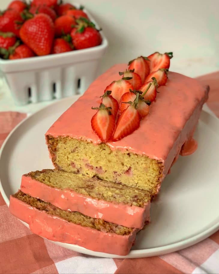 a loaf of strawberry bread with pink, strawberry frosting and sliced strawberries on top, with three slices cut into it.