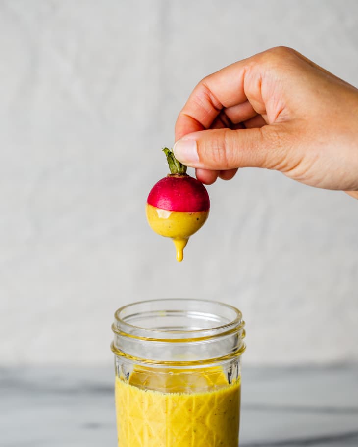 a hand holding a red radish by the greens, with half of it dipped into a pale yellow tahini dressing, and a small, glass jar with the rest of the dressing below.