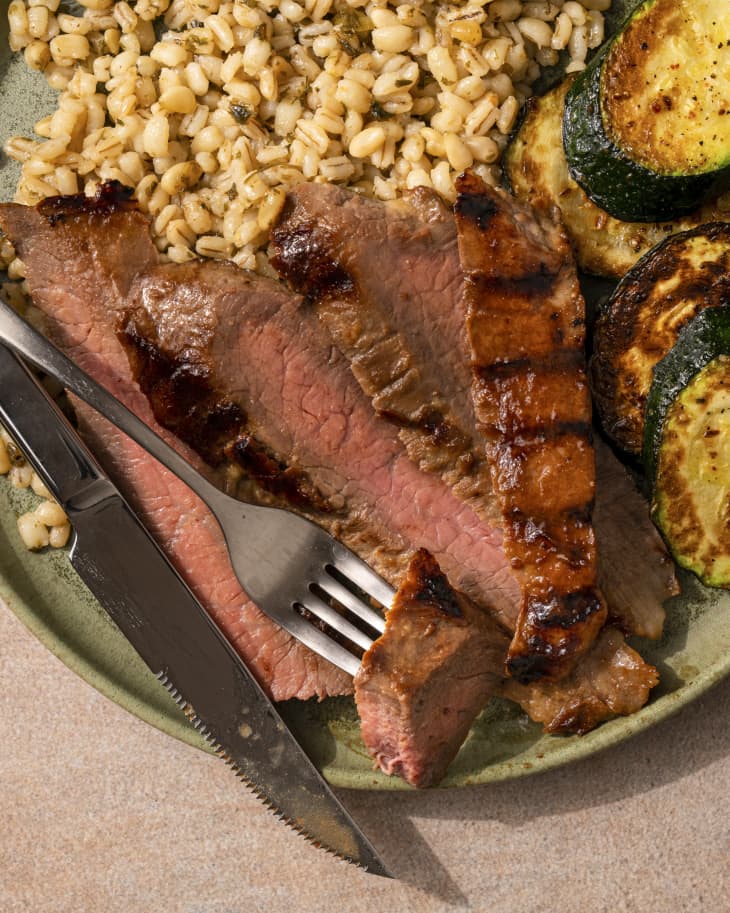 a closeup of medium rare, sliced, grilled, Flank steak, with a grain on the side, a grilled vegetable and fork and knife resting in the food.