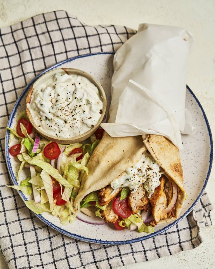 a chicken gyro on a plate, with a ramekin of tzatziki sauce and some shredded lettuce and diced tomatoes next to it.