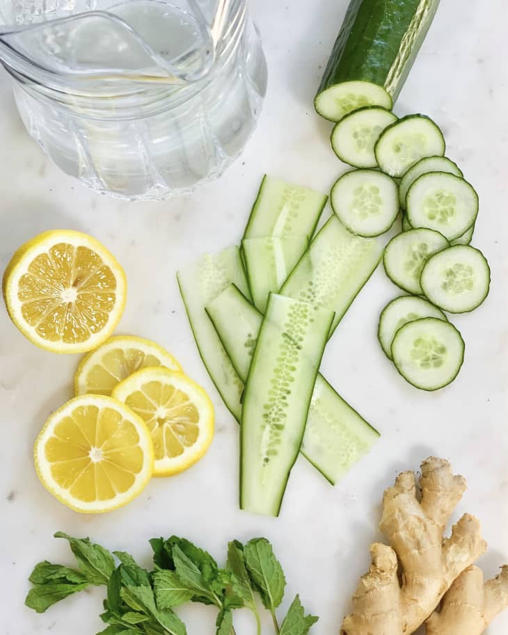 A photo of sliced cucumbers (cut both long ways and in rounds), sliced lemon rounds and fresh ginger next to a pitcher of water.