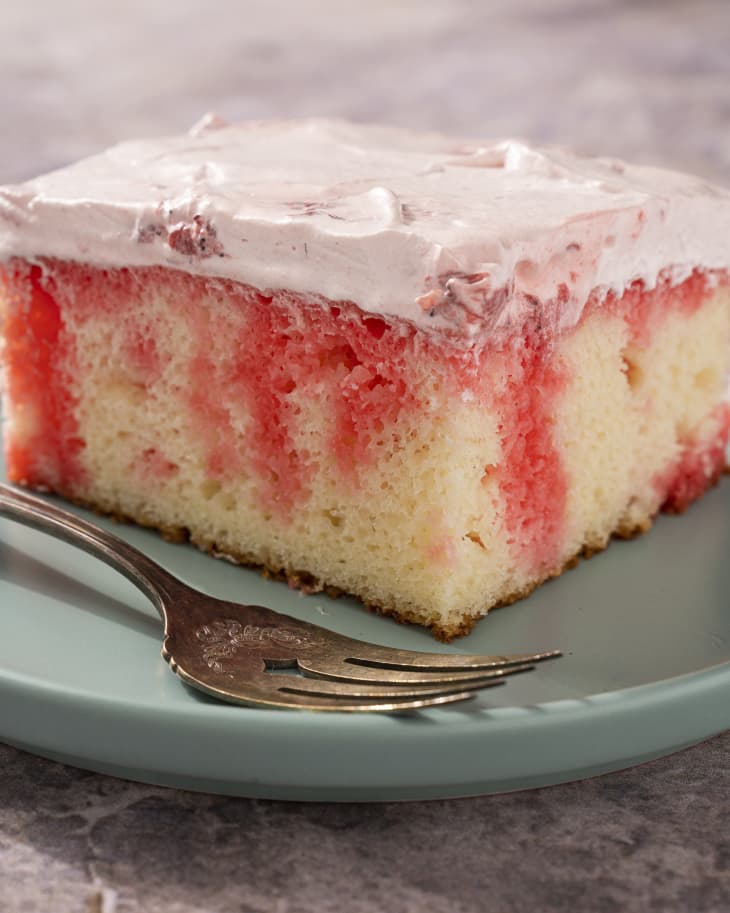 A piece of vanilla cake with jello drizzles poked throughout, topped with a smooth layer of whipped cream, with a fork on the side.