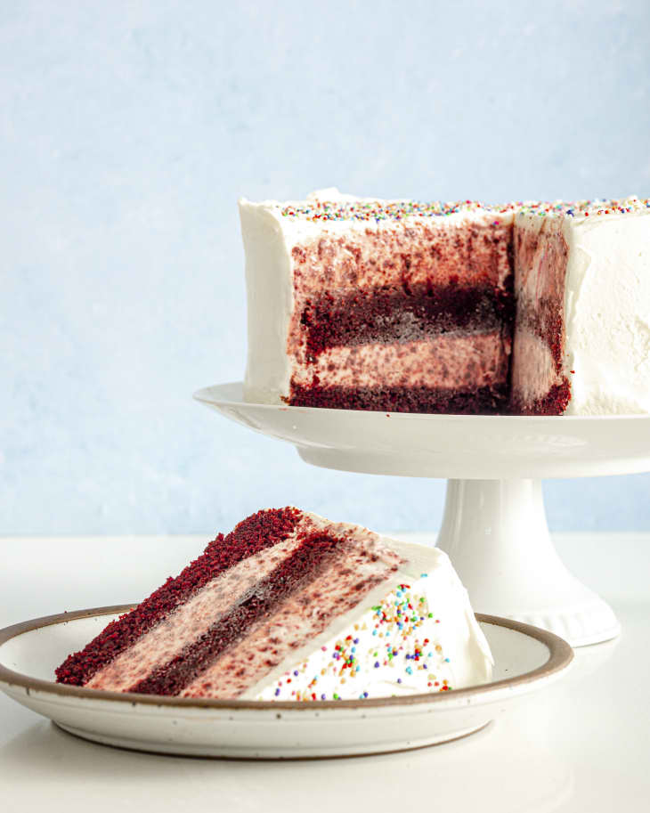 A photo of a slice of layered red velvet ice cream cake with the whole cake in the background
