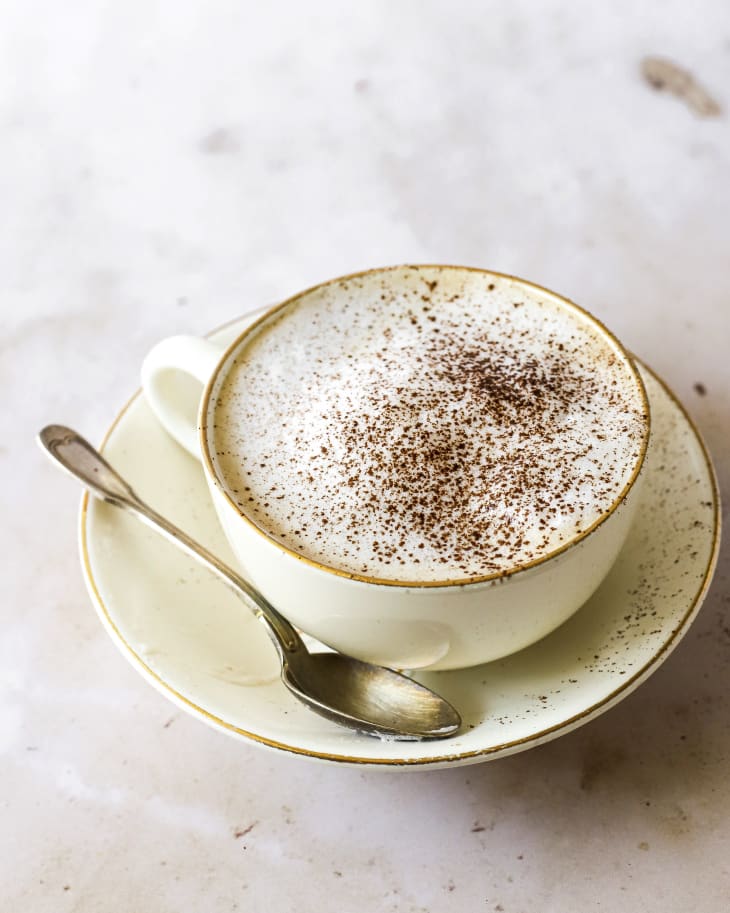 A photo of a cappuccino with cinnamon sprinkled on top, on a small saucer with a spoon on the side.