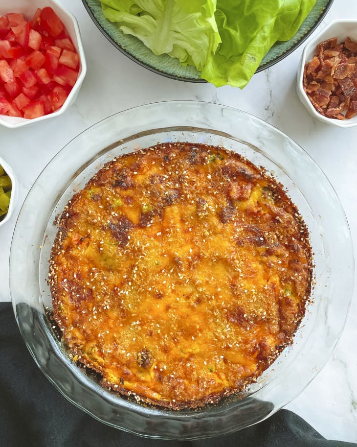 A photo of a cheeseburger pie, with melted cheese on top and some burger toppings (tomatoes, lettuce, and chopped bacon) in small bowls on the side.