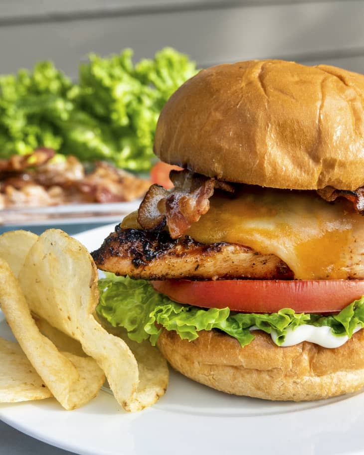 A photo of a grilled chicken sandwich on a bun with melted cheese, strips of bacon, lettuce, and a slice of tomato, with chips on the side. There is more food out of focus in the background.