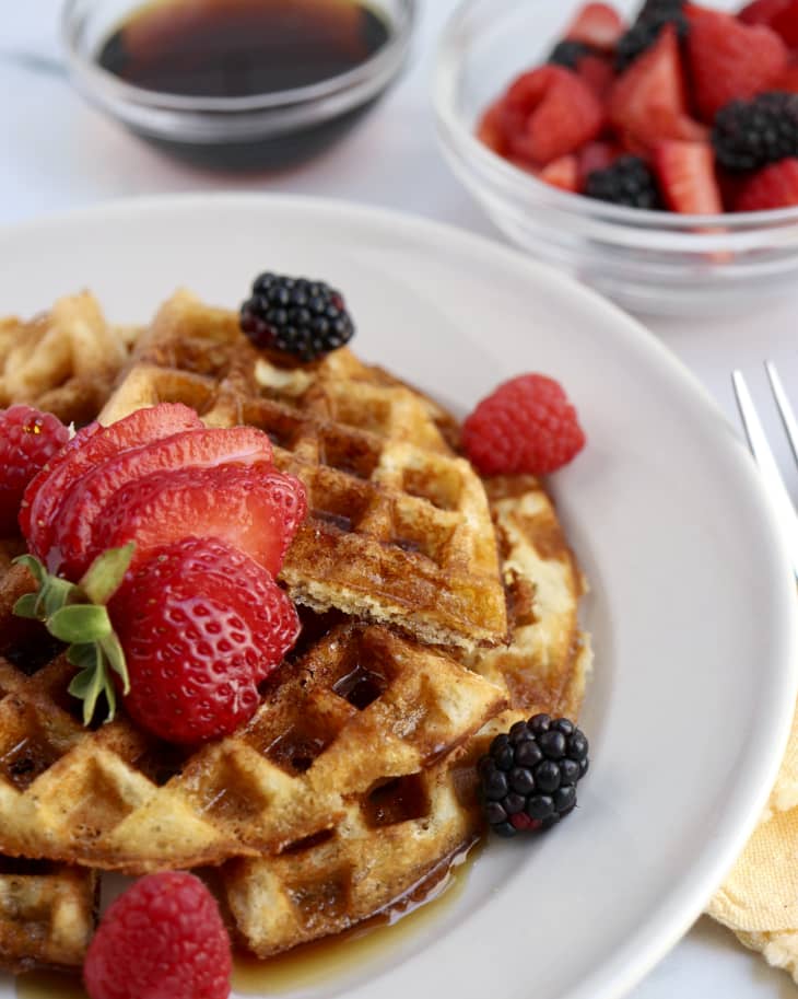 A photo of a plate of almond flour waffles with strawberries, raspberries and blackberries on top, and a small bowl of berries in the background.