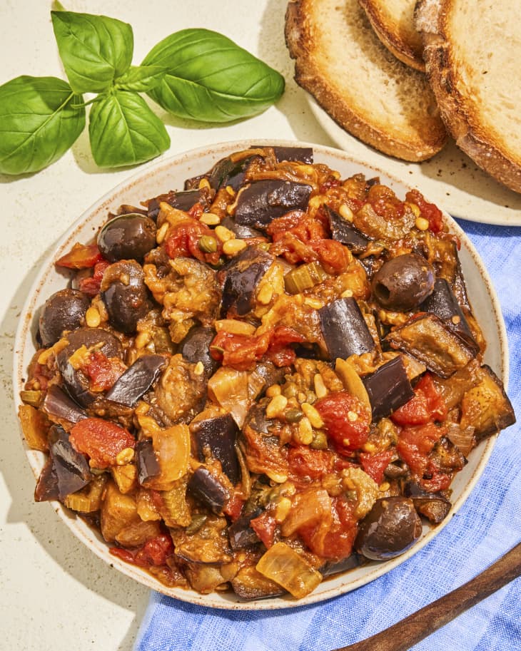 A photo of Caponata (chopped fried eggplant and other vegetables, seasoned with olive oil, tomato sauce, celery, olives, and capers) on a round plate with basil and sliced toast in the background.