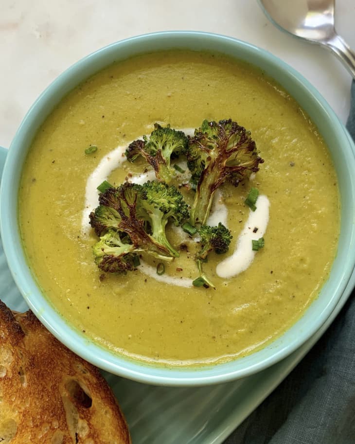 A photo of cream of broccoli soup in a turquoise bowl, garnished with cream and chives and roasted broccoli, with a piece of toast on the side.