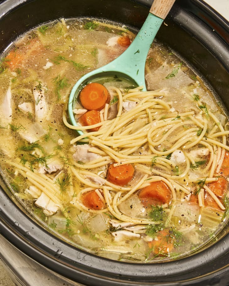 A photo of chicken noodle soup in a slow cooker, with a turquoise cooking spoon resting inside the slow cooker starting to lift up a spoonful.
