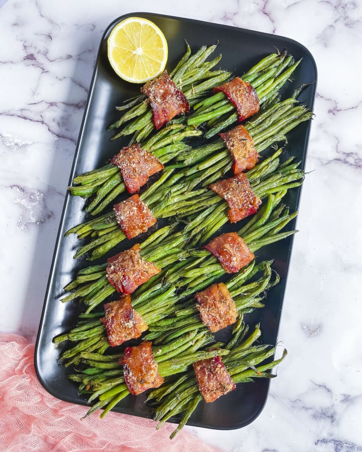 A photo of a tray of small bundles of cooked green beans, with each bundle wrapped in a slice of thick bacon.