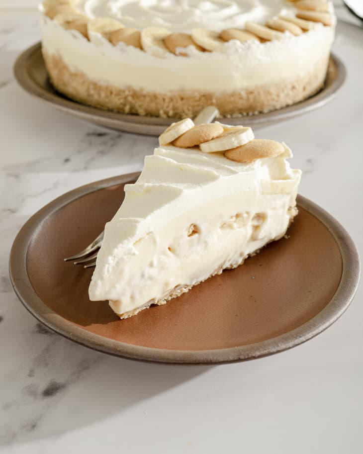 A photo of a slice of banana pudding cheesecake, with alternating slices of banana and nilla wafers around the rim of the cake, and the rest of the cake in the background.