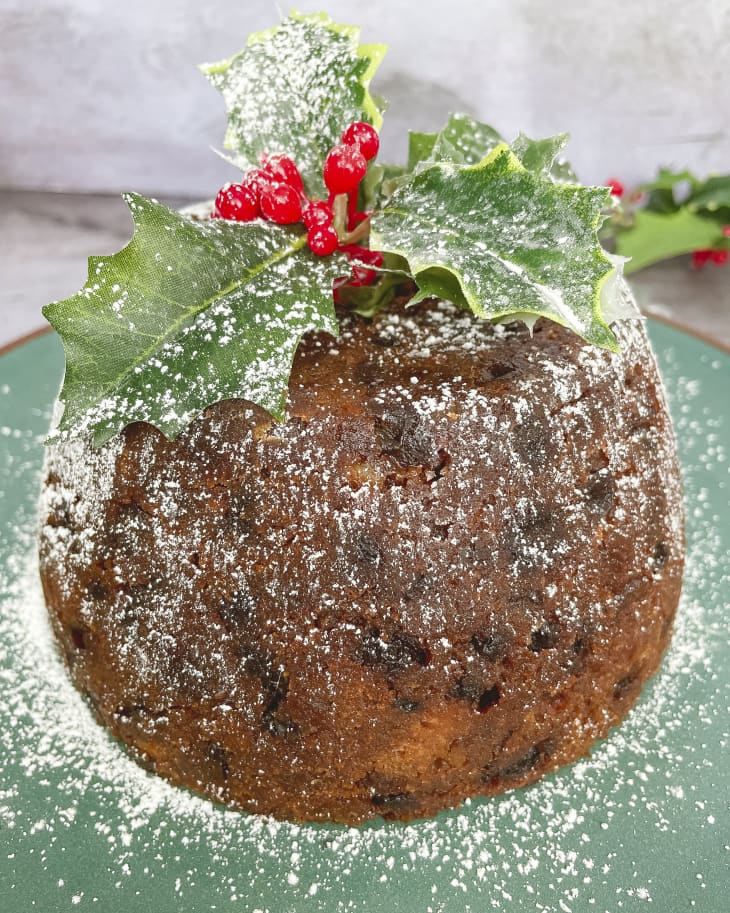 A photo of a whole Christmas Pudding/Figgy pudding (a traditional Christmas round cake made with dried fruit) topped with a holly sprig and powder sugar.