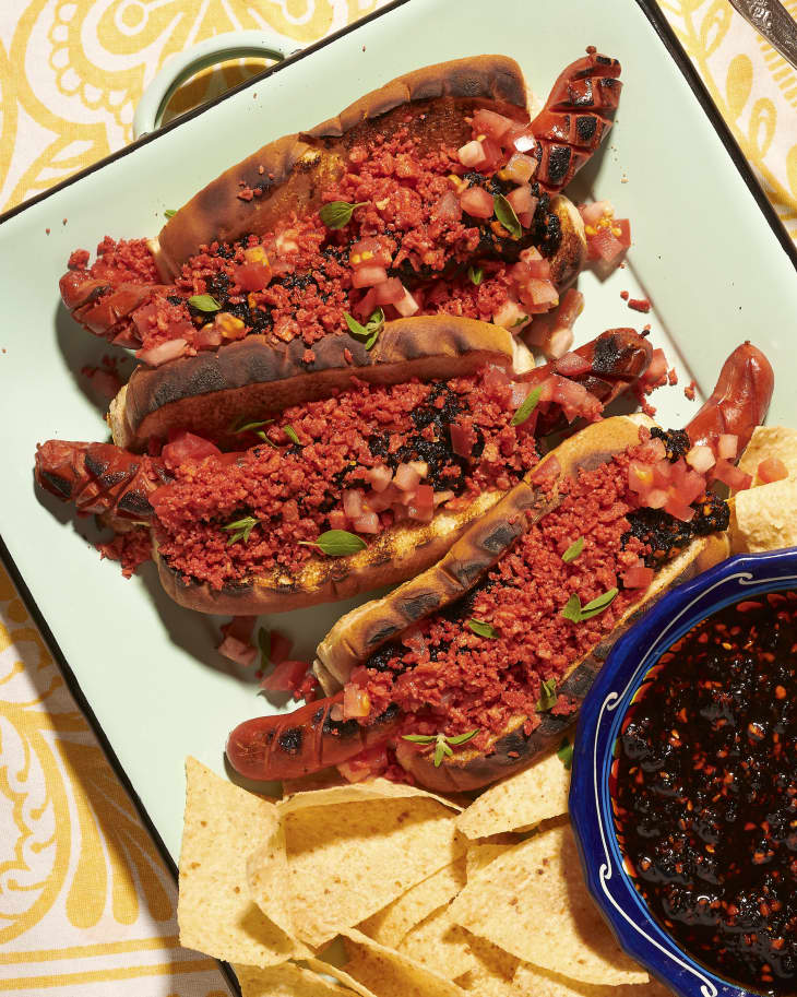 Grilled hot dogs with a red spice  topping, and chips and black bean dip on the side
