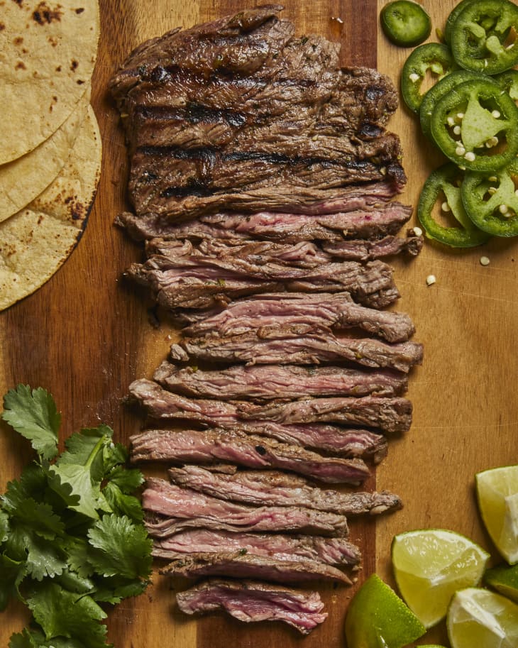 Carne Asada sliced on a cutting board with jalapeno peppers, lime wedges, cilantro, and corn tortillas