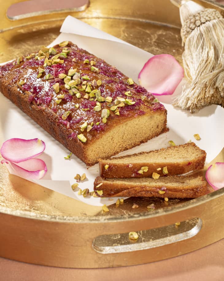Almond-tahini cake topped with rose syrup and pistachios, garnished with rose petals on a gold tray.