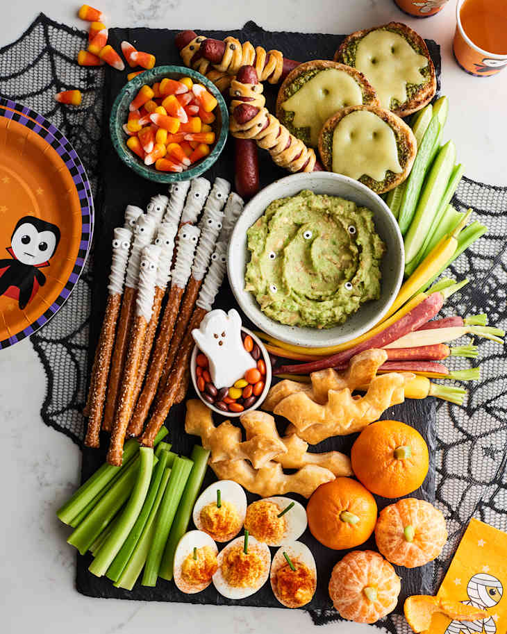 Festive Halloween snack board with mummy pretzels, guacamole, candy corn, and ghost-shaped cheese on crackers.