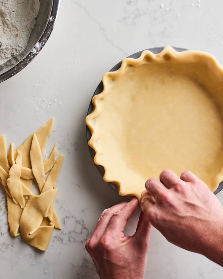 Overhead photo of hands crimping a pie crust. Strips of extra crust on the side, and a bowl of flour up in the corner of the frame.