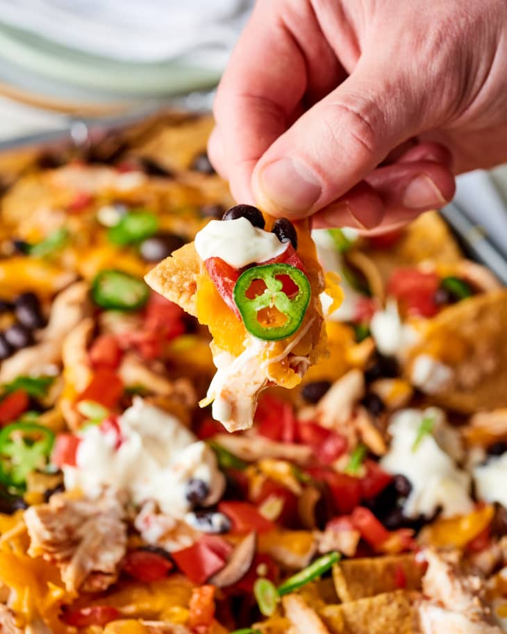 Close up of sheet pan chicken nachos, hand holds chip with cheese, chicken, sour cream, tomato, black olive and slice of jalapeno