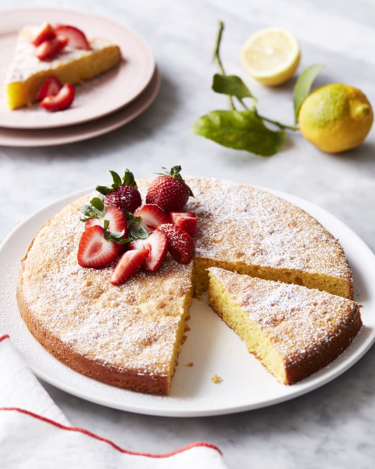 Whole flourless lemon almond cake with slice cut out. Topped with powered sugar and strawberries