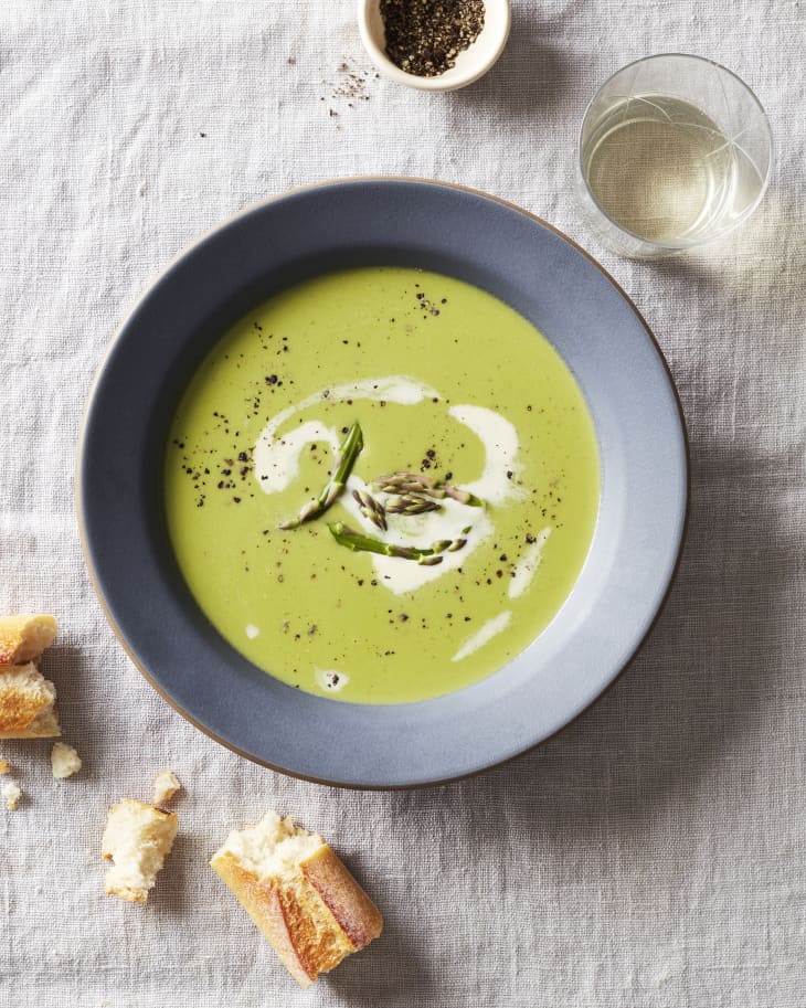 A bowl of cream of asparagus soup sits on a table next to a glass of white wine and bread.