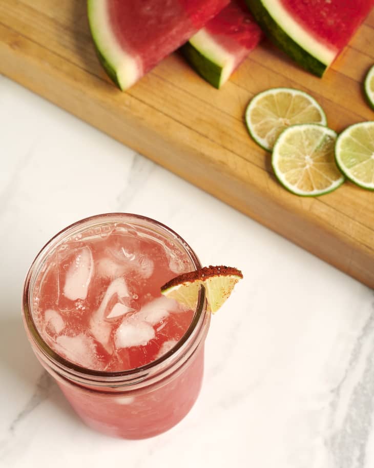 Cocktail garnished with lime sits on the kitchen counter next to a cutting board filled with watermelon wedges and limes.