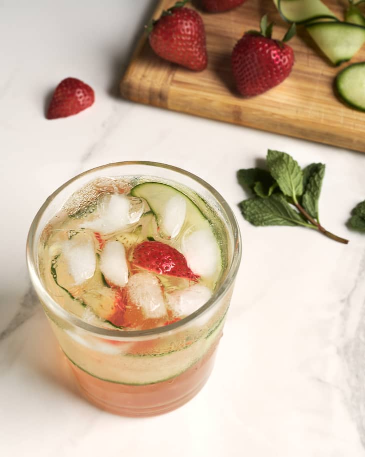 Cocktails garnished with cucumber and strawberries is sitting on a kitchen counter next to a cutting board filled with mint, strawberries, and cucumber.