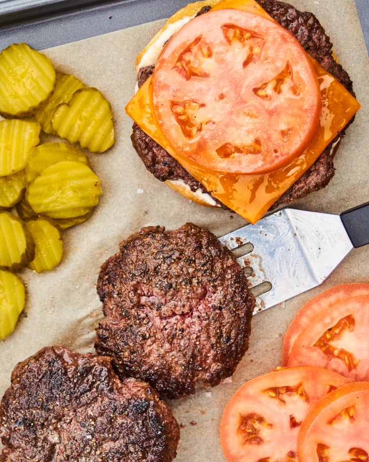 Burger patties with a partially assembled burger, pickles and sliced tomatoes