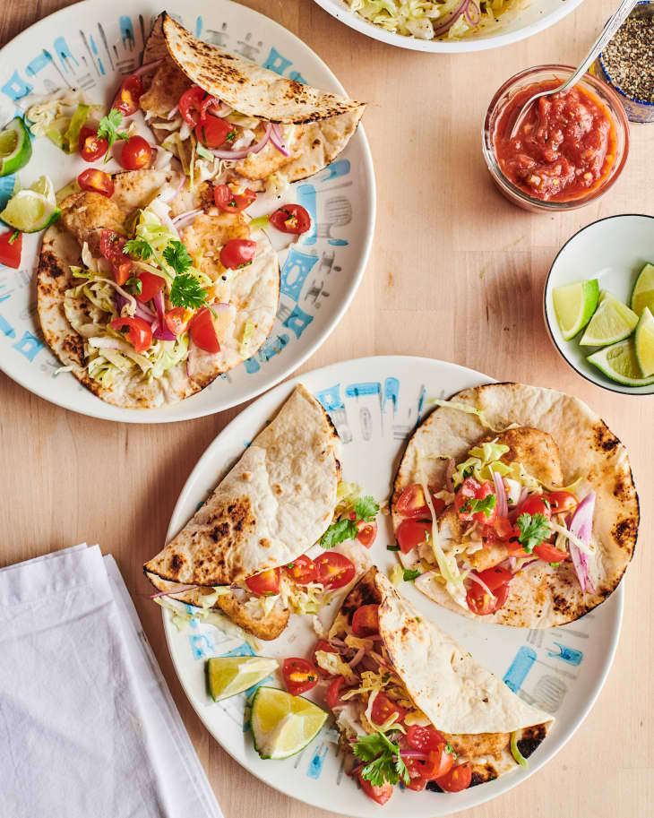 Five fish stick tacos sit on a  table next to quartered limes in a bowl, salsa, and pepper.