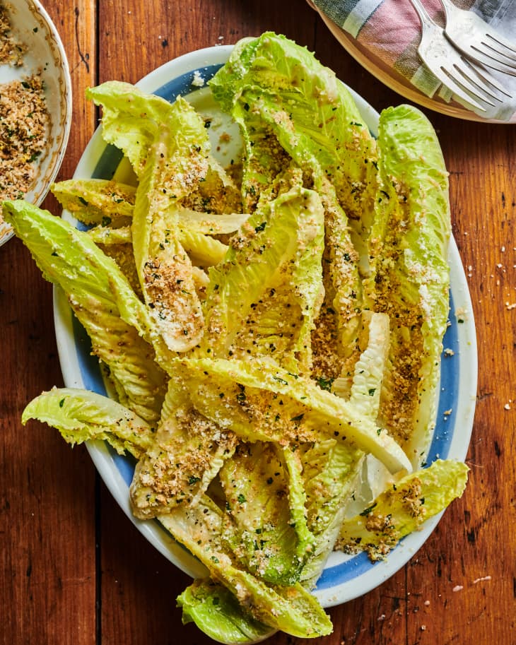 A platter of caesar salad sits on a wood table with a small bowl of breadcrumbs off to the side.