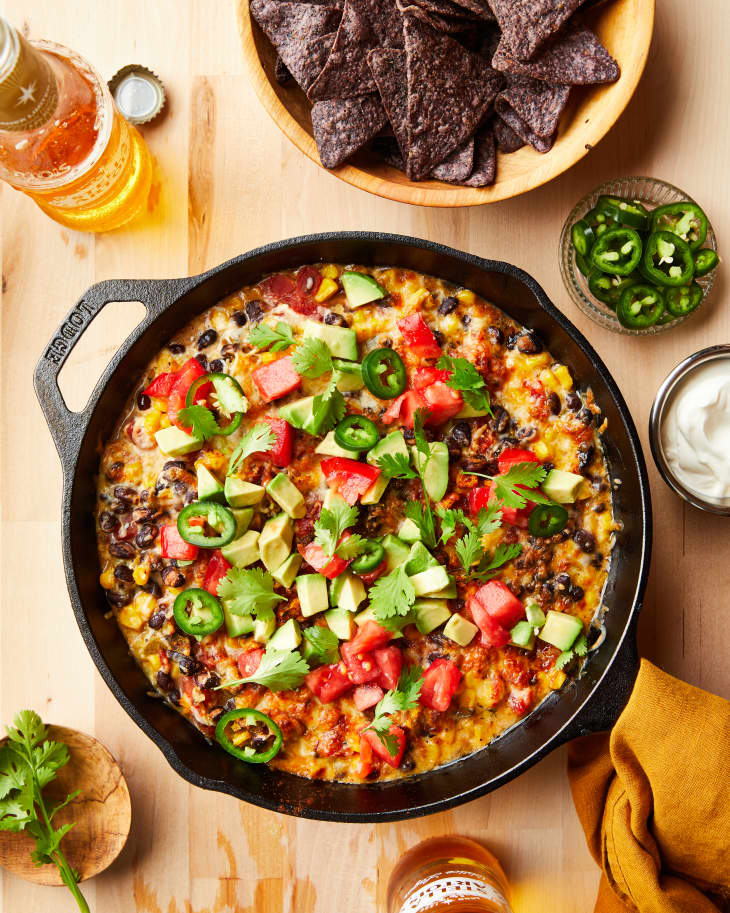 4 can pantry nacho dip in cast iron skillet surrounded by a bowl of blue corn tortilla chips, sliced jalapeño peppers, ramekin of sour cream, bottles of Mexican beer, a sprig of cilantro and mustard colored linen napkin.