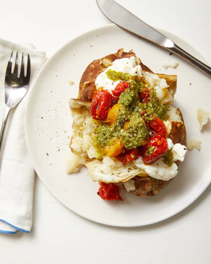Baked potato on a white plate, potato is topped with dollop of ricotta, blistered red and yellow cherry tomatoes and green pesto.